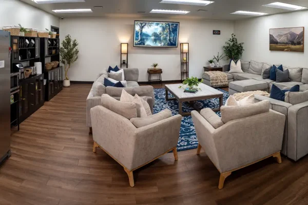 The men’s residential living room common area with comfortable couches and chairs, a TV, snacks, a toaster, airfryer, microwave, and fridge.